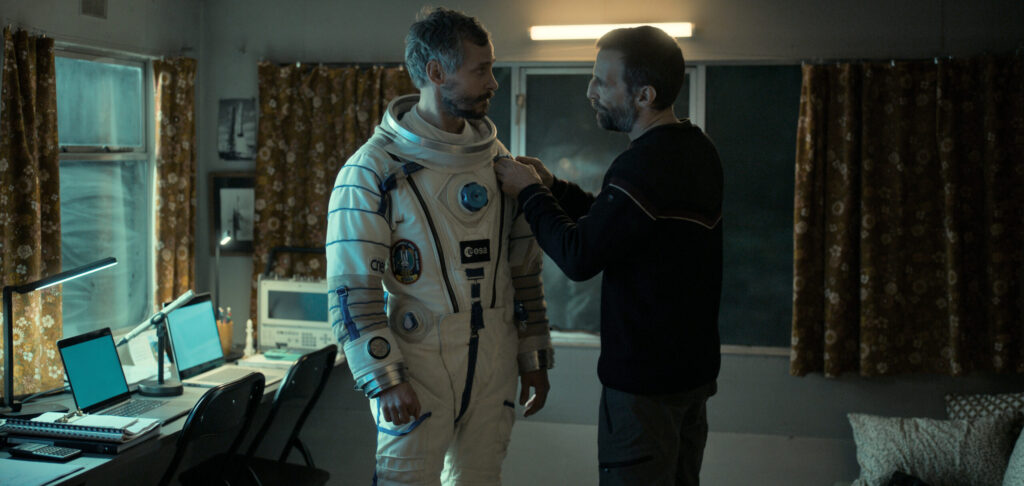 Homem com camisa preta ajeita traje aeroespacial de amigo, que está na sua frente