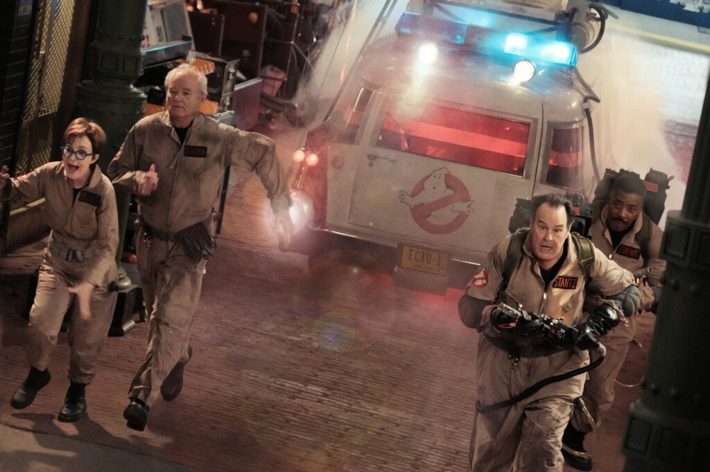 Elenco de Ghostbusters correndo com trajes e van atrás dele com logo da equipe